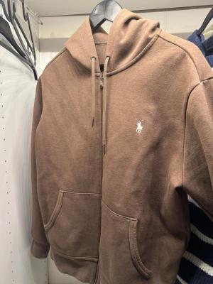 Brun hoodie från Polo Ralph Lauren - Snygg brun hoodie från Polo Ralph Lauren med dragkedja och huva. Tröjan har två stora fickor framtill och klassisk logga broderad i vitt på bröstet. Perfekt för chill dagar och enkel att matcha med jeans eller joggers.