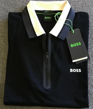 Svart pikétröja från BOSS med half zip - Svart pikétröja från BOSS med vit och mörkgrå krage, half zip framtill och BOSS-logga broderad på bröstet. Snygg och stilren design som passar perfekt till jeans eller chinos. Materialet känns mjukt och bekvämt mot huden. S-L storlek finns perfekt till Jul!🎄 