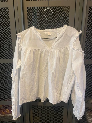 Vit blus med volang från Gina Tricot - Säljer en vit blus från Gina Tricot med v-ringning och långa ärmar. Blusen har volangdetaljer längs axlar och ärmar samt rynkad kant vid halsen. Tillverkad i mjuk bomull och har en luftig, loose passform. Perfekt för en chill och stilren look.