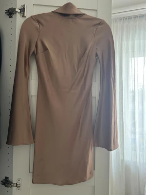 Champagnefärgad kort klänning Malina - Kort klänning från Malina i champagnefärgad satin. Klänningen har långa utsvängda ärmar och en bred, draperad krage i ryggen. Aldrig använd, ny utan prislapp.