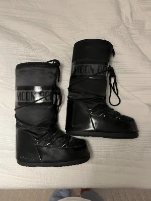 Svarta Moon Boots med snörning - Säljer ett par klassiska svarta Moon Boots med hög skaft och snörning framtill. Skorna har en glansig finish på nedre delen och tydlig logga runt skaftet. Perfekta för vintern och riktigt sköna att ha på sig när det är kallt ute.