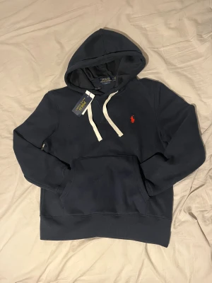 Helt ny! Mörkblå hoodie från Ralph Lauren - Snygg helt ny mörkblå hoodie från Polo Ralph Lauren i passformen slim fit med vit snörning och klassisk röd broderad logga på bröstet. Tröjan har huva, magficka och långa ärmar. Perfekt för dig som gillar stilrent och vill ha något bekvämt nu under vintern. Storlek: small. Nypris 2399kr, mitt pris 899kr