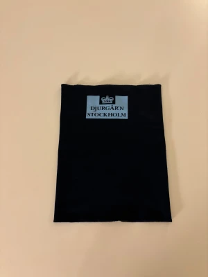 Svart halsduk Djurgår'n Stockholm - Svart tubhalsduk med tryckt ljusblå logga och texten 'Djurgår'n Stockholm' framtill. Halsduken är slät och har en synlig söm på baksidan. Perfekt för dig som vill visa ditt lagstöd och hålla dig varm.