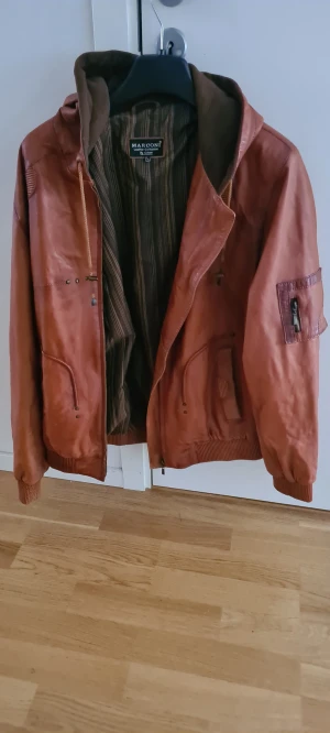 Brun skinnjacka från Marconi, strl L - Säljer en brun skinnjacka från Marconi Leather Collection i storlek L. Jackan har huva, ribbade muddar och flera fickor med dragkedja. Insidan är fodrad med ett brunt och gult randigt tyg. Perfekt för dig som vill ha en snygg och klassisk look.