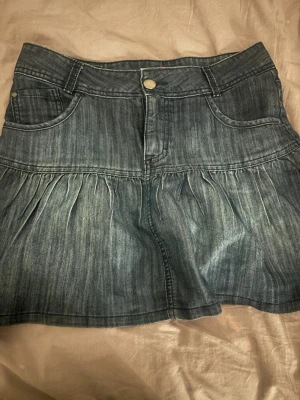 Mörkblå jeanskjol med fickor - Snygg mörkblå jeanskjol med klassisk femficksdesign och volangdetalj nedtill. Kjolen har knapp och dragkedja framtill samt bälteshällor. Perfekt för dig som gillar denim och vill ha en cool, avslappnad stil.