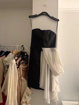 Bandå klänning - Snygg axelbandslös kortklänning i svart med en vit volangärm i transparent tyg. Klänningen har en figurnära passform.