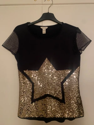 Svart topp med guldpaljetter från Monsoon, 12-13 år - Svart topp från Monsoon med korta transparenta ärmar i mesh och små prickar. Framsidan har en stor stjärna i guldiga paljetter. Strl 12-13 år. Materialet är mjukt och stretchigt.