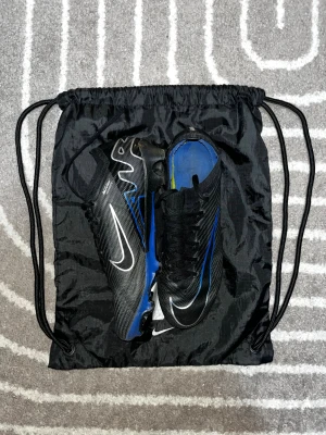Nike Mercurial fotbollsskor svart/blå - Säljer ett par Nike Mercurial fotbollsskor i svart med blå och vita detaljer. Skorna har snörning, platt sula med dobbar och är tillverkade i syntetmaterial. Perfekta för dig som vill ha lätta och snabba skor på planen. Snygg design med tydliga Nike-loggor.