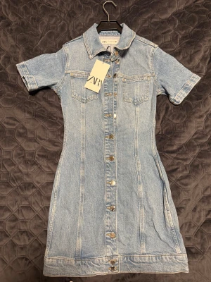 Jeansklänning med knappar från Zara - Snygg ljusblå jeansklänning från Zara med kort ärm, klassisk och krage och knappstängning hela vägen framtill. Klänningen har två bröstfickor och figurnära passform. Perfekt för dig som gillar en tidlös denimlook.