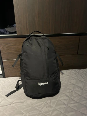 Svart ryggsäck från Supreme - Säljer en svart ryggsäck från Supreme med klassisk logga framtill och på axelremmarna. Väskan har flera fack, justerbara remmar och är tillverkad i slitstark polyester. Perfekt för skolan eller stan när du vill ha en clean och stilren look.