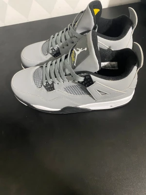 Nike Air Jordan 4 Retro Grå - Nike Air Jordan 4 Retro i grått med svarta och vita detaljer samt inslag av gult på tungan. Skorna har snörning, meshpaneler och den ikoniska Jumpman-loggan på både tunga och häl. Snygg och modern siluett med platt sula och rund tå, perfekt för dig som gillar streetwear.