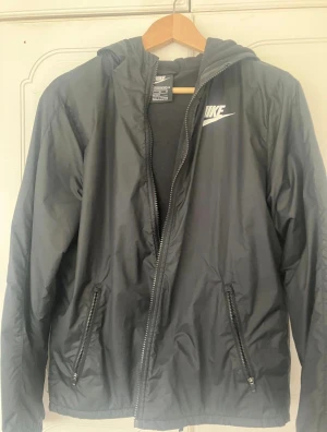 Svart vindjacka från Nike XL - Svart vindjacka från Nike i storlek XL med huva och dragkedja. Jackan har två fickor med dragkedjor och vit Nike-logga på bröstet. Insidan är fodrad med mjuk fleece och har en 'Just Do It'-detalj. Perfekt för blåsiga dagar.