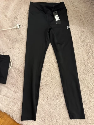 Svarta tights  - Svarta tights från  med hög midja och diskret logga på låret. Tillverkade i stretchigt syntetmaterial som sitter tight och bekvämt. Perfekta för träning eller chill dagar. Klassisk och stilren look.