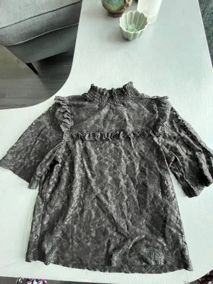 Svart spetsblus med volang från H&M Divided - Svart blus i spets från H&M Divided med hög krage och volangdetaljer över bröstet och axlarna. Blusen har halvkort ärm och är genombruten med blommigt spetsmönster. Perfekt för dig som gillar romantiska och trendiga detaljer. Pris går att diskutera 😊