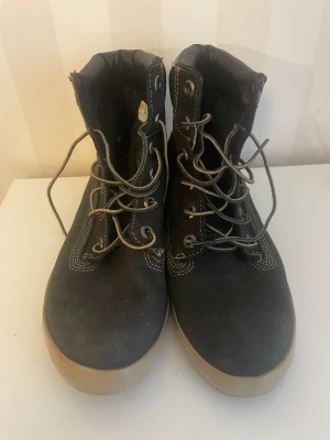 Svarta boots från Timberland st.38 - Svarta boots från Timberland med beige sula och kontrasterande vita sömmar. Skorna har snörning med metallhål och är tillverkade i mocka och skinn. Perfekta för höst och vinter med sin robusta design och vadderad kant upptill.