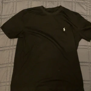 Svart t-shirt från Polo Ralph Lauren (prisförlsag) - Svart t-shirt från Polo Ralph Lauren med klassisk rund hals och korta ärmar. Diskret vit Polo-logga broderad på bröstet. Tillverkad i mjuk bomull för skön passform och stilren look. Perfekt till jeans eller shorts.