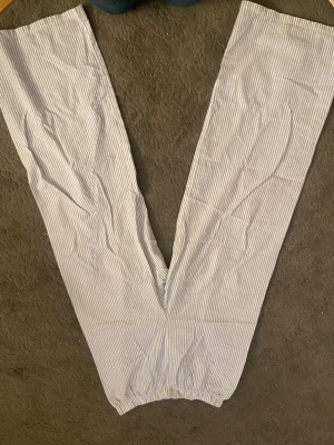 Blå vitt pyjamasset från H&M - Säljer ett par ljusblå och vita pyjamasbyxor från H&M i storlek M med tillhörande pyjamaslinne med blåa band och en blå blomma på bröstet. Byxorna har smala vertikala ränder, elastisk midja och är tillverkade i mjuk bomull. Perfekta för chill hemma eller mysiga kvällar.