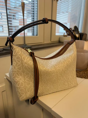 Vit/Beige väska  - Snygg axelväska i vit canvas med broderat mönster och detaljer i brunt skinn. Väskan har en bred axelrem i brunt läder, guldfärgad dragkedja och en stabil botten i skinn. Perfekt storlek för det viktigaste och riktigt trendig design. Skriv för mer info 