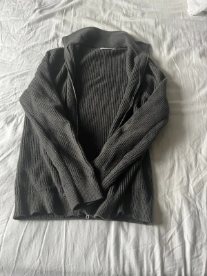 Mörkgrå ribbad kofta med dragkedja från East West - Snygg mörkgrå kofta med ribbad struktur och dragkedja framtill. Perfekt för lager-på-lager och har en hög krage som ger extra stilpoäng. Materialet är stickat och känns mjukt mot huden. Passar dig som gillar en clean och enkel look. Nypris 1000 knappt använd