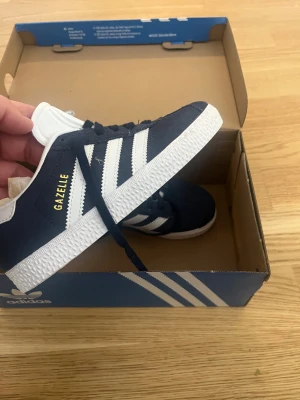 Nya Adidas Gazelle mörkblå sneakers strl 33 - Säljer ett par klassiska Adidas Gazelle sneakers i mörkblå mocka med vita detaljer och de ikoniska tre ränderna på sidorna. Skorna har snörning, vit plös med Adidas-logga och platt vit sula. Perfekta för dig som gillar retrostil och streetwear.