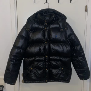 Svart dunjacka från Polo Ralph Lauren - Säljer en svart, glansig dunjacka från Polo Ralph Lauren. Jackan har en pufferdesign med långa ärmar och en praktisk huva. Den är utrustad med både dragkedja och knappar för extra skydd mot kyla. jackan är i barnstorlek storlek xl. Passar mig hyfsat som har M och är 180 cm lång. Lite liten, kort i ärmarna. Har en liten defekt på vänster armen. Det är ett hål som har limmats ihop och gjorts svart . Syns knappt vid användning ! Se sista bilden. Tveka inte att höra av er vid frågor.