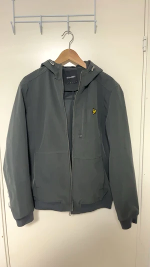Grå vindjacka från Lyle & Scott  -  Ny pris koster 1650kr,Snygg grå vindjacka från Lyle & Scott med huva och dragkedja framtill. Jackan har det klassiska gula fågel-loggan på bröstet, två stora fickor och ribbade muddar. Perfekt för blåsiga dagar och enkel att matcha med det mesta.