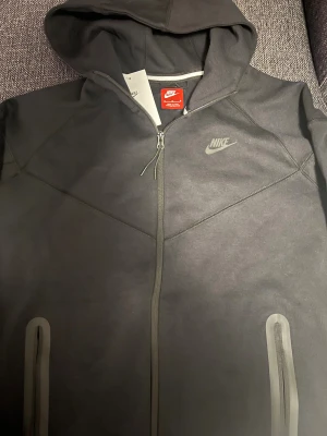 Svart Nike tech fleece - Svart hoodiejacka från Nike med hel dragkedja, huva och två fickor med dragkedja framtill. Jackan har en stilren look med Nike-logga på bröstet och är tillverkad i mjukt bomullsmaterial. Perfekt för en sportig och avslappnad stil. Sitter som en L även