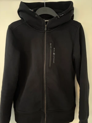 Svart hoodie från Sail Racing M - Svart hoodie från Sail Racing i storlek M. Tröjan har huva med snörning, hel dragkedja framtill och en bröstficka med zip. Insidan är fodrad med mjuk fleece för extra värme. Snyggt broderad logga på huvan och stilren design. Liten i storleken.