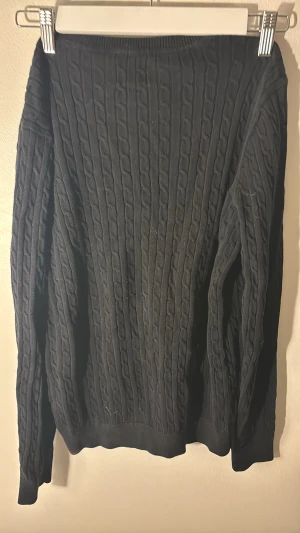 Svart kabelstickad tröja Crocker M - Svart kabelstickad tröja från Crocker i storlek M. Tröjan har långärmade ärmar, ribbade muddar och klassisk rund hals. Materialet är mjukt och stickat, perfekt för kyliga dagar. Snygg struktur med kabelmönster som ger en cool och tidlös vibe. Finns liten missfärgning vid nederkant på tröjan men väldigt litet som man knappt lägger märke till.