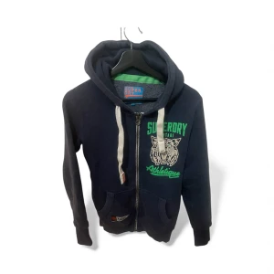 Mörkblå hoodie från Superdry XS - Snygg mörkblå hoodie från Superdry med grönt och vitt tryck fram och bak. Tröjan har huva med vita snören, dragkedja och två fickor framtill.