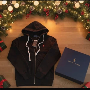 Svart hoodie från Polo Ralph Lauren - Svart hoodie från Polo Ralph Lauren med vit snörning och dragkedja framtill. Tröjan har huva, långärmade ärmar och ett litet rött broderat logomärke på bröstet. Insidan är mjuk och fleecefodrad för extra komfort. Perfekt för kalla dagar.