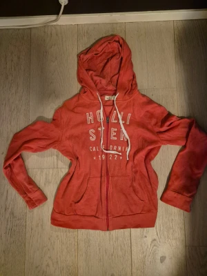 Röd hoodie från Hollister med dragkedja - Säljer en röd hoodie från Hollister med vit text på bröstet och snörning i huvan. Tröjan har lång ärm, fickor framtill och dragkedja. 