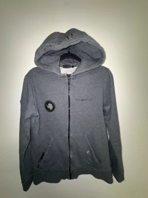 Grå hoodie från Sail Racing - Snygg grå hoodie från Sail Racing med svart tryck 'Antarctica Expedition' på ryggen och logga på bröstet. Tröjan har hel dragkedja, huva och två fickor framtill. Perfekt för chill dagar och har en clean look med sportig vibe. Märke vid dragkedja och ficka 
