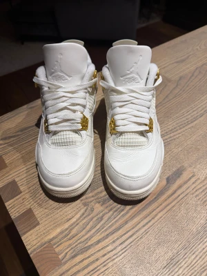 Nike Air Jordan 4 vit/guld sneakers - Nike Air Jordan 4 sneakers i helvit med snygga gulddetaljer på snörningen. Skorna har en klassisk siluett med rund tå, platt sula och meshpaneler på sidorna. Tillverkade i skinn med vadderad insida för extra komfort. Perfekt för dig som gillar stilrena och exklusiva sneakers.