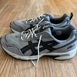 Grå Asics 1090v2 sneakers - Säljer ett par grå Asics 1090v2 sneakers med svarta detaljer och vit sula. Skorna har mesh- och mockapaneler, klassisk snörning och rund tå. Perfekta för dig som gillar sportig stil och bekväma skor till vardags.