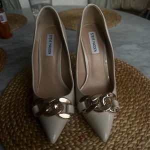 Beige pumps från Steve Madden - Snygga pumps i beige lack från Steve Madden med spetsig tå och hög klack. Framtill sitter en chunky guldfärgad kedjedetalj som ger skorna en lyxig vibe. Perfekta för dig som vill sticka ut med elegans och stil.