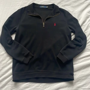 Ralph Lauren Half Zip - Svart långärmad tröja från Polo Ralph Lauren med klassisk röd broderad logga på bröstet. Tröjan har en snygg half zip-dragkedja vid halsen och är tillverkad i mjukt bomullsmaterial. Perfekt för en clean och stilren look.