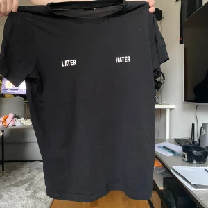 Svart t-shirt med textprint - Svart t-shirt med vit text 'LATER' och 'HATER' tryckt på bröstet. Klassisk rund hals och korta ärmar. Enkel och stilren design som passar perfekt till jeans eller shorts. Materialet känns mjukt och bekvämt mot huden.