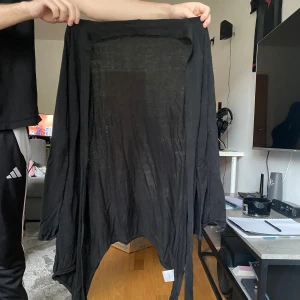 Svart tunn kofta med knytband - Svart, supertunn och luftig kofta med långa ärmar och knytband framtill. Perfekt att slänga över en topp för en chill och avslappnad look. Asymmetrisk nederkant ger en cool vibe.