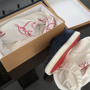 Mörkblå sneakers från Louboutin - Säljer ett par mörkblå sneakers från Louboutin med klassisk röd sula och coola nitar på hälen. Skorna är i mocka och har vita sulor. Perfekta för dig som vill sticka ut med en lyxig och edgy stil.