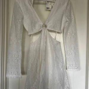 Vit spetsklänning med ringdetalj - Vit kortklänning med spetsmönster och långärmade ärmar. Klänningen har en ringdetalj framtill och öppet parti vid midjan. Aldrig använd. 