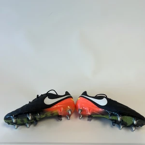 Nike Tiempo fotbollsskor svart/orange - Nike Tiempo fotbollsskor med svart ovandel i skinn, vita Nike loggor och orange detaljer vid hälen. Skorna har snörning och metall-dobbar för extra grepp på planen. Snygg design med inslag av gult och grönt på sulan, perfekt för dig som vill sticka ut på matchen.