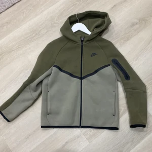 Nike grön vindjacka med huva XL - Nike vindjacka i två gröna nyanser med huva och hel dragkedja framtill. Jackan har en cool svart detalj med dragkedja på ena ärmen och svarta kantband. Perfekt för dig som gillar sportig stil och vill ha något som sticker ut.