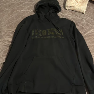 Svart hoodie från BOSS med guldtryck - Svart hoodie från BOSS med stort guldigt logotryck framtill. Klassisk huva med snörning och känguruficka. Tillverkad i mjuk polyesterblandning som känns skön mot huden. Perfekt för dig som vill ha en stilren och trendig look.