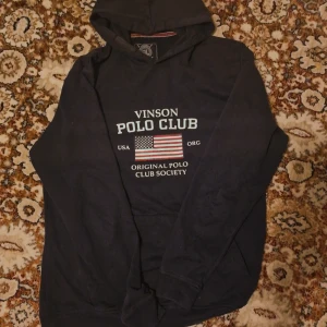 Svart hoodie från Vinson Polo Club -  Använd ett par gånger helt utan fläck eller smuts. svart hoodie från Vinson Polo Club med tryck på bröstet och amerikansk flagga. Tröjan har huva och långa ärmar, samt en klassisk känguruficka framtill. Materialet är en skön blandning av bomull och polyester, perfekt för chill dagar.