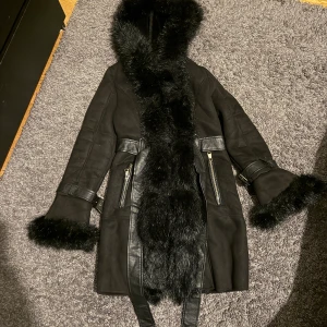 Svart pälsjacka med huva och bälte - Svart lång pälsjacka med fluffig fuskpäls längs huva, ärmslut och framkant. Jackan har bälte i midjan, dragkedjor på fickorna och snygga detaljer i skinnimitation. Perfekt för kalla dagar när du vill sticka ut lite extra.
