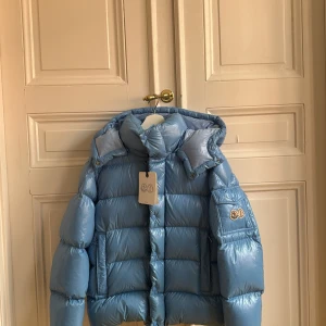 Moncler Maya 70th anniversary  - Säljer en blå pufferjacka från Moncler med glansig finish och huva. Jackan har Moncler-logga på ärmen, praktiska fickor och dragkedja framtill. Fodret är mjukt och jackan är perfekt för kalla dagar. Snygg, modern look med quiltad design och coola detaljer. Är i mycket gott skick men där hangarn tyvärr släppt! Kvitto och tags medföljer 