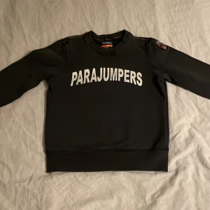 Svart fleece-tröja Parajumpers - Svart fleece-tröja från Parajumpers med stor vit logga på bröstet. Tröjan har långärmade ärmar, ribbade muddar och rund hals. Märkespatch på axeln och orange detaljer vid halsen. Perfekt för dig som gillar streetwear och snygga loggor.