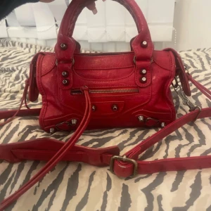 Röd handväska i skinn - Cool röd handväska i skinn med silverfärgade nitar och detaljer. Väskan har både handtag och justerbar axelrem, samt dragkedja framtill och på sidan. Perfekt storlek för det viktigaste och ger en edgy vibe till din outfit.