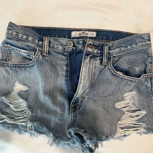 Blå jeansshorts med slitningar Hollister - Snygga blå jeansshorts från Hollister med råa, fransiga kanter och slitna detaljer framtill. Klassisk femficksmodell med knapp och dragkedja. 